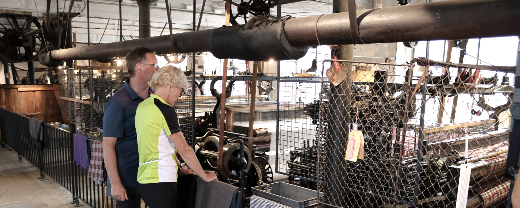 Zwei Personen stehen in einer historischen Textilfabrik und betrachten alte Maschinen. Stoffproben hängen am Geländer., © Nordeifel Tourismus GmbH Zwei Personen stehen in einer historischen Textilfabrik und betrachten alte Maschinen. Stoffproben hängen am Geländer., © Nordeifel Tourismus GmbH