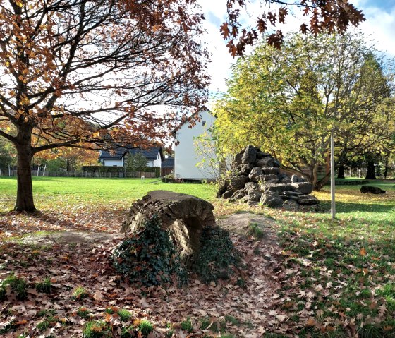 Roman water pipe in Nettersheim, surrounded by autumnal trees and foliage. Houses and a blue sky can be seen in the background., © Seco GmbH Roman water pipe in Nettersheim, surrounded by autumnal trees and foliage. Houses and a blue sky can be seen in the background., © Seco GmbH