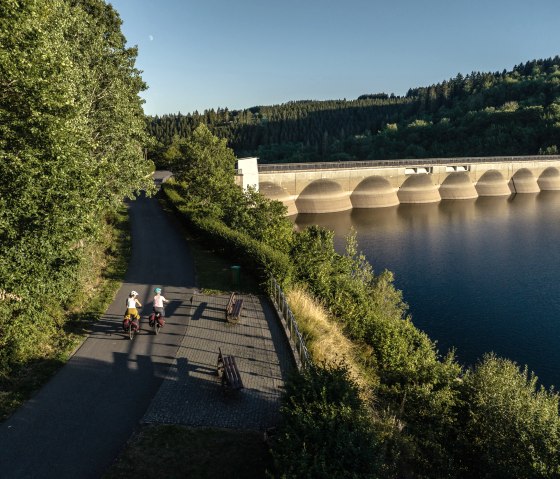 Zwei Radfahrer fahren auf einem Weg entlang der Oleftalsperre. Die Staumauer und der See sind von Bäumen umgeben, bei klarem Himmel und Sonnenschein., © Eifel Tourismus GmbH, Dennis Stratmann Zwei Radfahrer fahren auf einem Weg entlang der Oleftalsperre. Die Staumauer und der See sind von Bäumen umgeben, bei klarem Himmel und Sonnenschein., © Eifel Tourismus GmbH, Dennis Stratmann