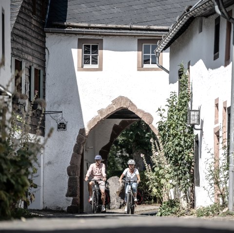 Zwei Radfahrer mit Helm fahren durch den historischen Burgort Kronenburg durch eine Häusergruppe hindurch., © Eifel Tourismus GmbH, Dennis Stratmann Zwei Radfahrer mit Helm fahren durch den historischen Burgort Kronenburg durch eine Häusergruppe hindurch., © Eifel Tourismus GmbH, Dennis Stratmann