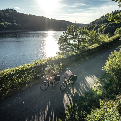Two cyclists ride along a lake in the sunshine, surrounded by green nature. The sun is reflected in the water., © Eifel Tourismus GmbH, Dennis Stratmann Two cyclists ride along a lake in the sunshine, surrounded by green nature. The sun is reflected in the water., © Eifel Tourismus GmbH, Dennis Stratmann