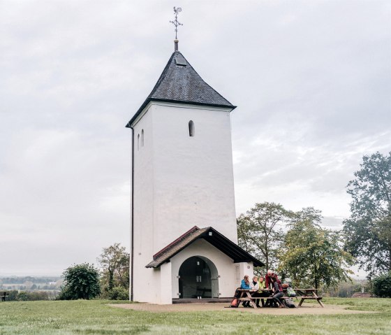 Weißer Turm mit spitzem Dach auf einer Wiese, Menschen sitzen an einem Picknicktisch. Bewölkter Himmel, umgeben von Bäumen., © Paul Meixner Weißer Turm mit spitzem Dach auf einer Wiese, Menschen sitzen an einem Picknicktisch. Bewölkter Himmel, umgeben von Bäumen., © Paul Meixner