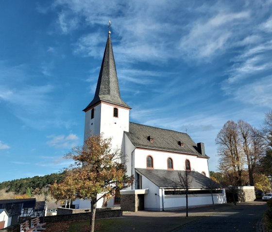 The church of St. Martin in Nettersheim against a blue sky, surrounded by autumnal trees and a cobbled path., © Sweco GmbH The church of St. Martin in Nettersheim against a blue sky, surrounded by autumnal trees and a cobbled path., © Sweco GmbH