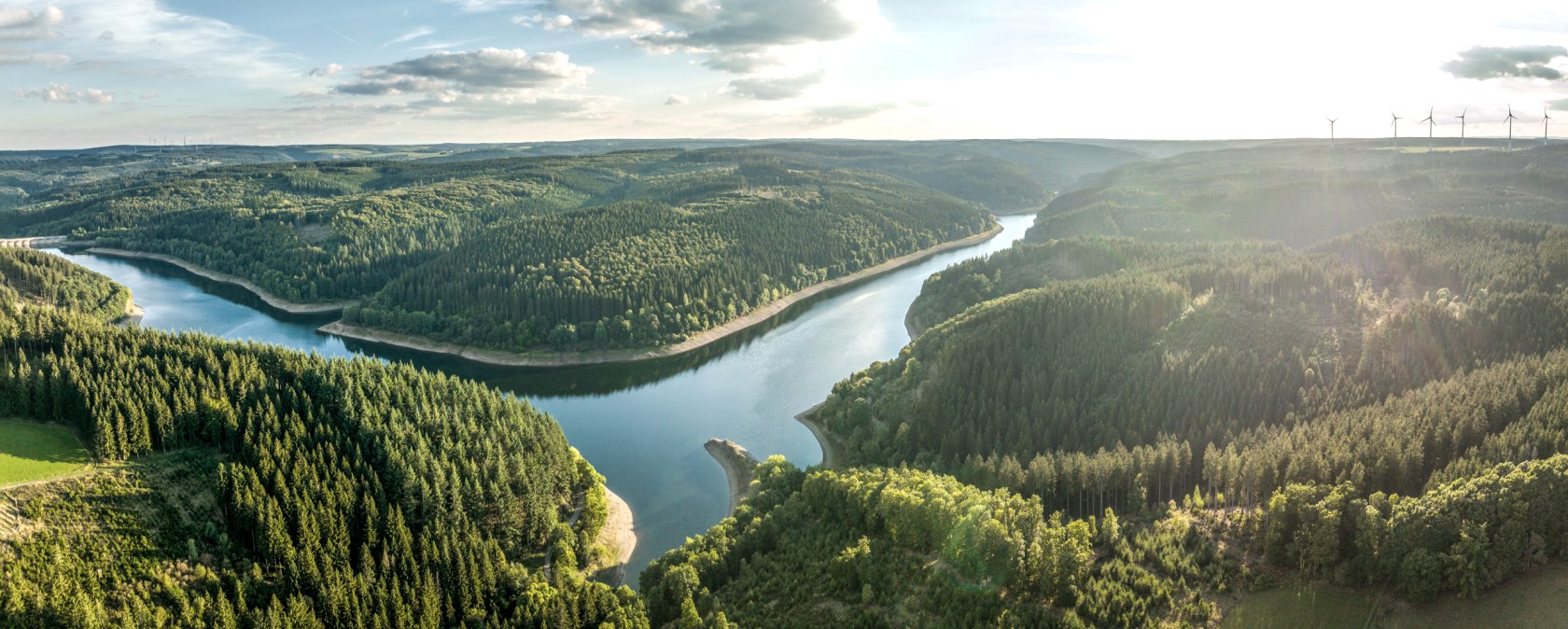 Aerial view of the Oleftalsperre, surrounded by dense forests. Wind turbines can be seen in the background against a sunny sky., © Eifel Tourismus GmbH, Dominik Ketz - finanziert durch REACT-EU Aerial view of the Oleftalsperre, surrounded by dense forests. Wind turbines can be seen in the background against a sunny sky., © Eifel Tourismus GmbH, Dominik Ketz - finanziert durch REACT-EU