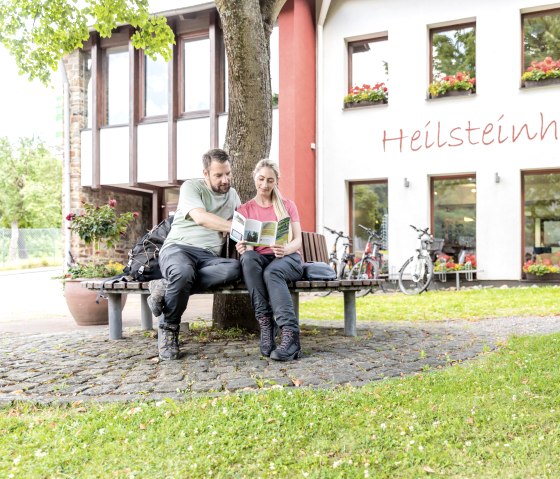 Two people are sitting on a bench in front of the Heilsteinhaus in Einruhr, reading a brochure. Bicycles are parked in front of the building, which is decorated with flowers., © -@Eifel Tourismus GmbH, AR-shapefruit-AG Two people are sitting on a bench in front of the Heilsteinhaus in Einruhr, reading a brochure. Bicycles are parked in front of the building, which is decorated with flowers., © -@Eifel Tourismus GmbH, AR-shapefruit-AG
