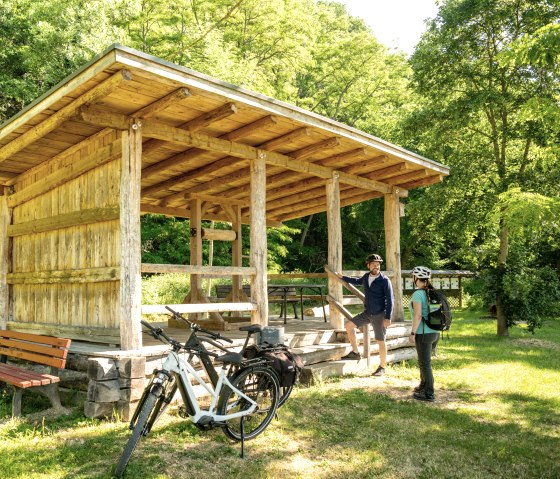 Zwei Radfahrer mit Helmen stehen neben einem hölzernen Unterstand im Grünen. Fahrräder sind abgestellt, umgeben von Bäumen und Wiese., © Eifel Tourismus GmbH, Dominik Ketz Zwei Radfahrer mit Helmen stehen neben einem hölzernen Unterstand im Grünen. Fahrräder sind abgestellt, umgeben von Bäumen und Wiese., © Eifel Tourismus GmbH, Dominik Ketz