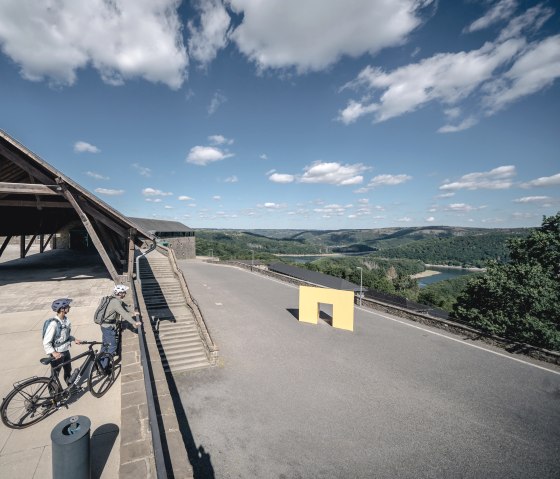 Two cyclists stand in front of a building with a wooden roof and look out over a hilly landscape with a river. The sky is blue with a few clouds., © Eifel Tourismus GmbH, Dennis Stratmann Two cyclists stand in front of a building with a wooden roof and look out over a hilly landscape with a river. The sky is blue with a few clouds., © Eifel Tourismus GmbH, Dennis Stratmann