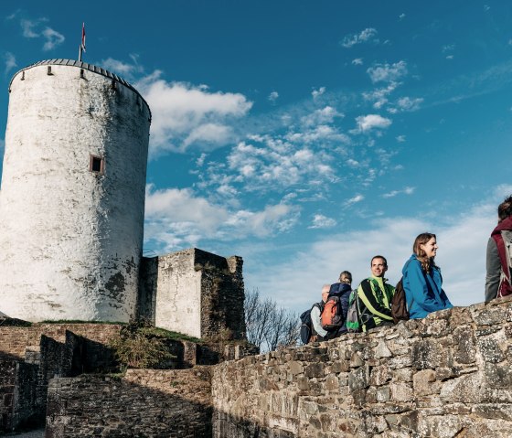 Menschen sitzen auf einer Steinmauer bei Burg Reifferscheid. Ein runder Turm mit Fahne ist zu sehen. Der Himmel ist blau mit wenigen Wolken., © Paul Meixner Menschen sitzen auf einer Steinmauer bei Burg Reifferscheid. Ein runder Turm mit Fahne ist zu sehen. Der Himmel ist blau mit wenigen Wolken., © Paul Meixner