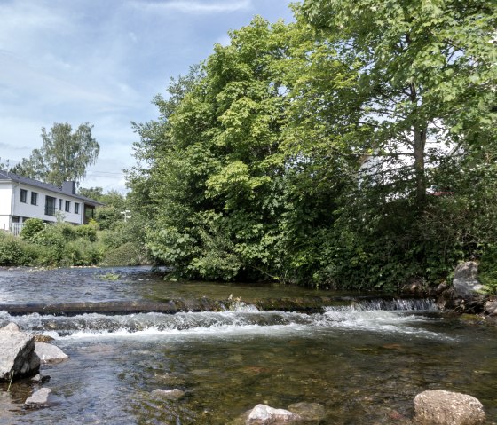 Kleiner Fluss mit flachem Wasser und Steinen, umgeben von grünen Bäumen und einem weißen Haus im Hintergrund., © Nordeifel Tourismus Kleiner Fluss mit flachem Wasser und Steinen, umgeben von grünen Bäumen und einem weißen Haus im Hintergrund., © Nordeifel Tourismus