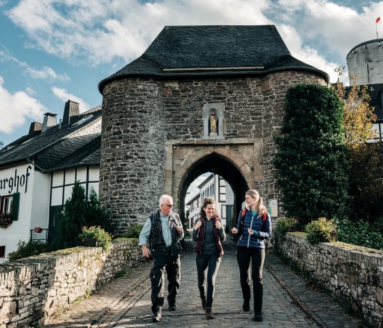 Drei Personen spazieren durch ein historisches Tor in Reifferscheid. Im Hintergrund sind Fachwerkhäuser und ein Turm zu sehen., © Paul Meixner Drei Personen spazieren durch ein historisches Tor in Reifferscheid. Im Hintergrund sind Fachwerkhäuser und ein Turm zu sehen., © Paul Meixner