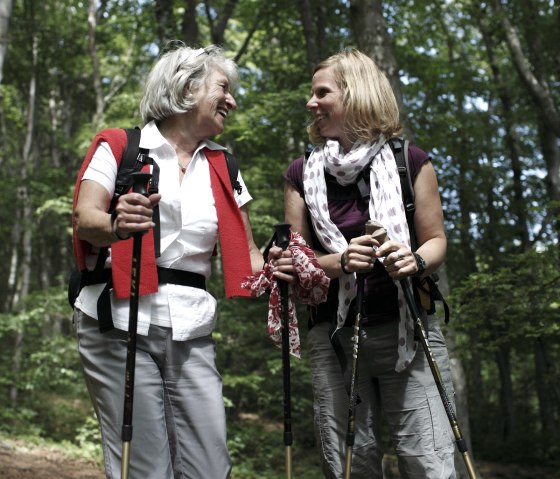 Two women are walking in the forest, smiling and equipped with walking sticks. They are wearing light clothing and enjoying nature., © Wandermagazin/ N. Glatter Two women are walking in the forest, smiling and equipped with walking sticks. They are wearing light clothing and enjoying nature., © Wandermagazin/ N. Glatter