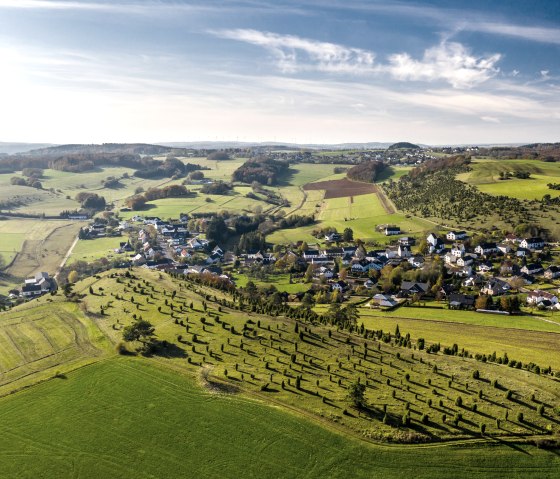 Luftaufnahme einer grünen Hügellandschaft mit einem kleinen Dorf in der Eifel. Felder und Bäume prägen die Umgebung unter einem blauen Himmel., © Eifel Tourismus, D. Ketz Luftaufnahme einer grünen Hügellandschaft mit einem kleinen Dorf in der Eifel. Felder und Bäume prägen die Umgebung unter einem blauen Himmel., © Eifel Tourismus, D. Ketz