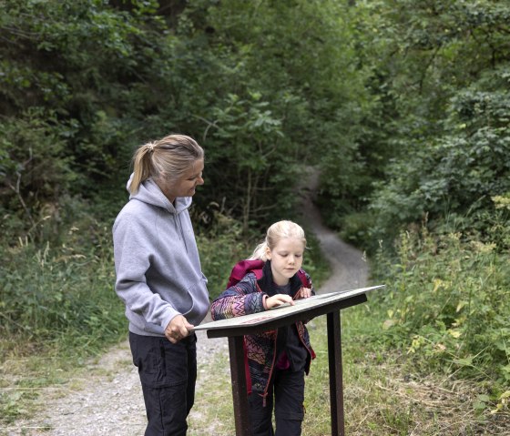 Eine Frau und ein Kind stehen im Wald und lesen gemeinsam eine Infotafel. Der Weg ist von üppigem Grün umgeben., © Eifel Tourismus, Tobias Vollmer Eine Frau und ein Kind stehen im Wald und lesen gemeinsam eine Infotafel. Der Weg ist von üppigem Grün umgeben., © Eifel Tourismus, Tobias Vollmer