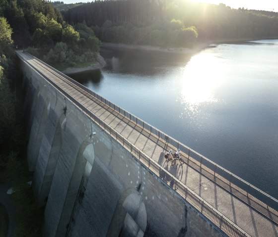 Luftaufnahme der Oleftalsperre in der Eifel. Die Staumauer verläuft diagonal, Radfahrer auf der Mauer, Wasser reflektiert Sonnenlicht., © Eifel Tourismus GmbH, Dennis Stratmann Luftaufnahme der Oleftalsperre in der Eifel. Die Staumauer verläuft diagonal, Radfahrer auf der Mauer, Wasser reflektiert Sonnenlicht., © Eifel Tourismus GmbH, Dennis Stratmann