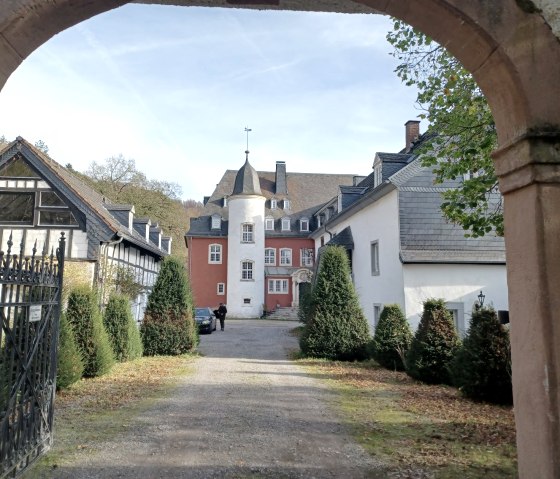 Blick durch ein Tor auf die Burg Dalbenden. Das Hauptgebäude ist von Bäumen umgeben, mit einem roten und weißen Turm in der Mitte., © Sweco GmbH Blick durch ein Tor auf die Burg Dalbenden. Das Hauptgebäude ist von Bäumen umgeben, mit einem roten und weißen Turm in der Mitte., © Sweco GmbH