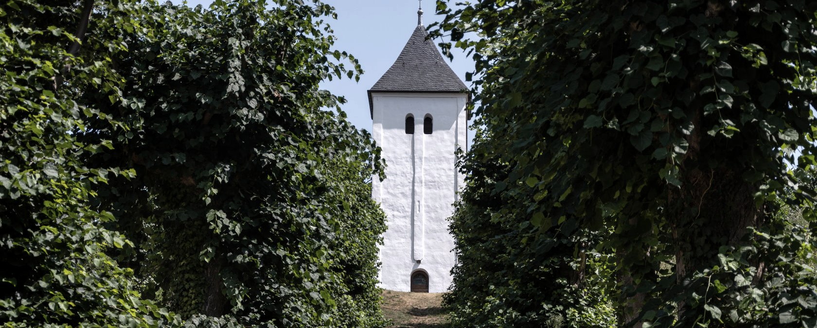 Ein weißer Turm steht am Ende einer Allee, die von hohen, grünen Bäumen gesäumt ist. Der Himmel ist klar und blau., © Nordeifel Tourismus GmbH Ein weißer Turm steht am Ende einer Allee, die von hohen, grünen Bäumen gesäumt ist. Der Himmel ist klar und blau., © Nordeifel Tourismus GmbH