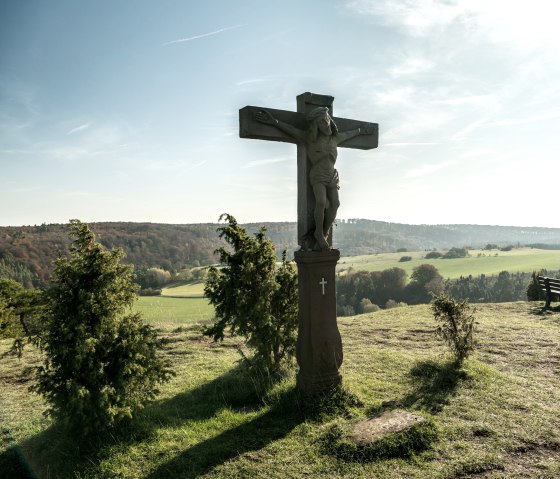 Ein Kreuz auf einem Hügel mit einer Bank daneben, umgeben von grüner Landschaft und bewaldeten Hügeln unter blauem Himmel., © Eifel Tourismus, D. Ketz Ein Kreuz auf einem Hügel mit einer Bank daneben, umgeben von grüner Landschaft und bewaldeten Hügeln unter blauem Himmel., © Eifel Tourismus, D. Ketz