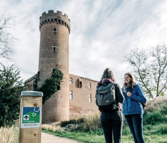 Zwei Personen vor einem historischen Turm in Zülpich. Ein Wegweiser der Eifelspur steht im Vordergrund. Der Himmel ist bewölkt., © Paul Meixner Zwei Personen vor einem historischen Turm in Zülpich. Ein Wegweiser der Eifelspur steht im Vordergrund. Der Himmel ist bewölkt., © Paul Meixner