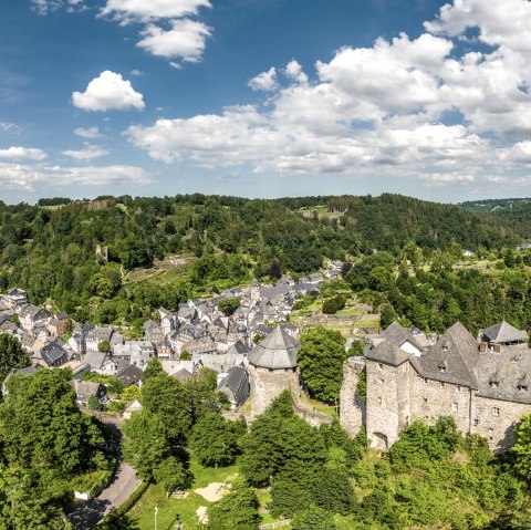 Panoramablick auf Monschau mit der Burg im Vordergrund, umgeben von grüner Landschaft und bewaldeten Hügeln unter blauem Himmel mit Wolken., © Eifel Tourismus GmbH, Dominik Ketz Panoramablick auf Monschau mit der Burg im Vordergrund, umgeben von grüner Landschaft und bewaldeten Hügeln unter blauem Himmel mit Wolken., © Eifel Tourismus GmbH, Dominik Ketz