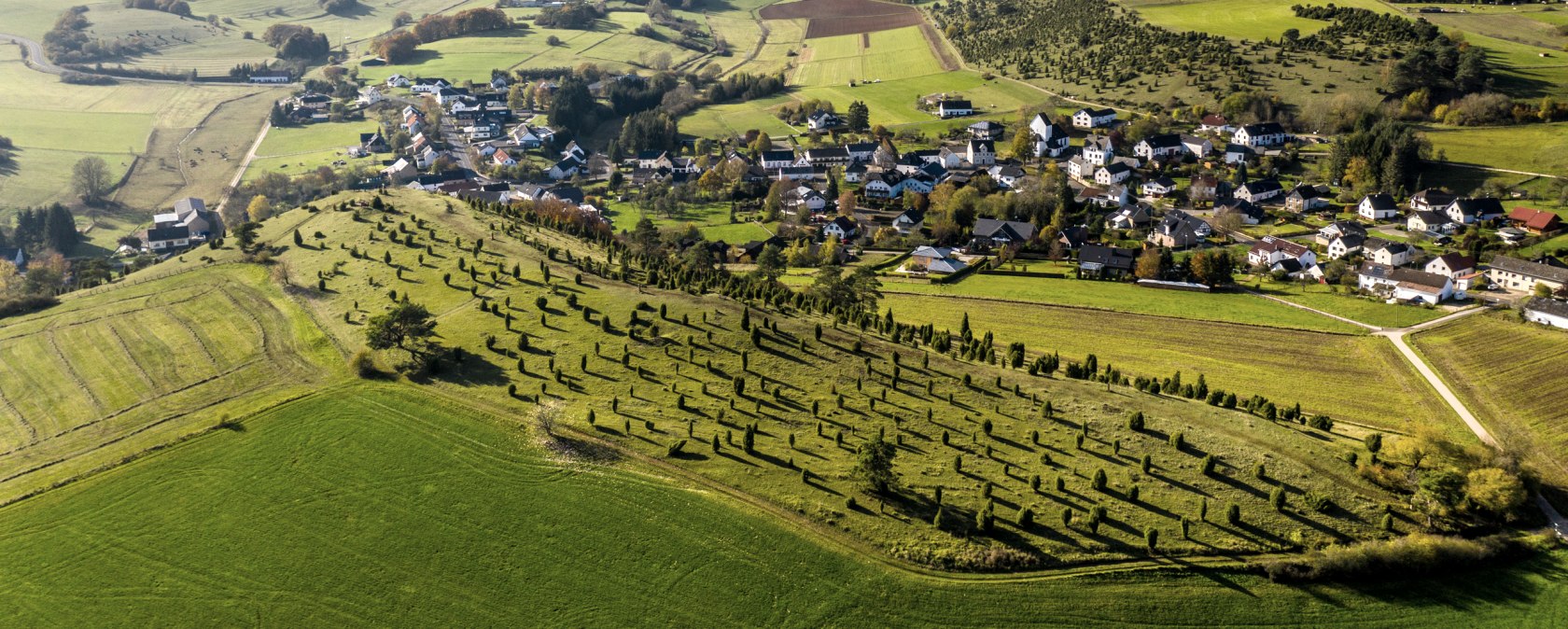 Luftaufnahme einer grünen Hügellandschaft mit einem kleinen Dorf in der Eifel. Felder und Bäume prägen die Umgebung unter einem blauen Himmel., © Eifel Tourismus, D. Ketz Luftaufnahme einer grünen Hügellandschaft mit einem kleinen Dorf in der Eifel. Felder und Bäume prägen die Umgebung unter einem blauen Himmel., © Eifel Tourismus, D. Ketz