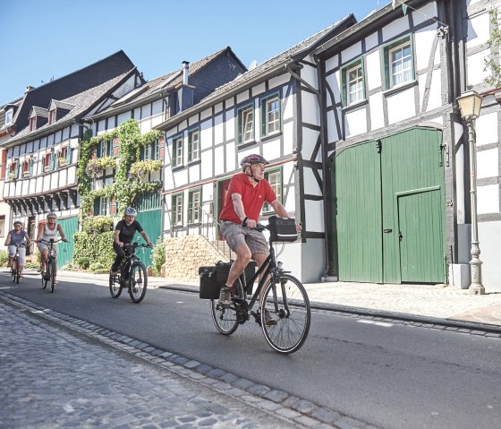 Cyclists ride through a street of half-timbered houses in the historic town center of Kommern. The sun is shining and the houses are decorated with plants., © Alexander Pallmer Cyclists ride through a street of half-timbered houses in the historic town center of Kommern. The sun is shining and the houses are decorated with plants., © Alexander Pallmer