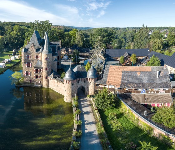 Aerial view of Satzvey Castle with moat, surrounded by green landscape and buildings. Tents can be seen on a meadow next to the castle., © Paul Meixner Aerial view of Satzvey Castle with moat, surrounded by green landscape and buildings. Tents can be seen on a meadow next to the castle., © Paul Meixner