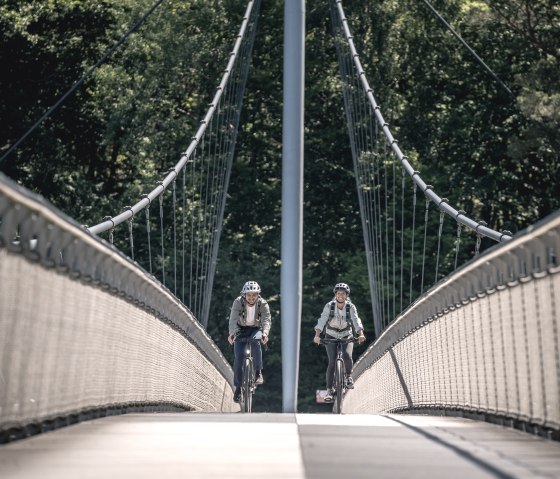 Two cyclists cross the Victor Neels Bridge, a steel suspension bridge surrounded by dense forest., © Eifel Tourismus GmbH, Dennis Stratmann Two cyclists cross the Victor Neels Bridge, a steel suspension bridge surrounded by dense forest., © Eifel Tourismus GmbH, Dennis Stratmann