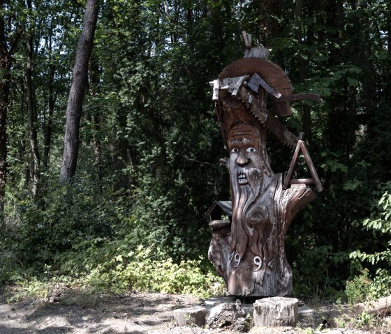 Holzskulptur eines Mannes mit Säge im Wald, umgeben von Bäumen und Pflanzen. Die Skulptur trägt die Jahreszahl 1994., © Nordeifel Tourismus GmbH Holzskulptur eines Mannes mit Säge im Wald, umgeben von Bäumen und Pflanzen. Die Skulptur trägt die Jahreszahl 1994., © Nordeifel Tourismus GmbH