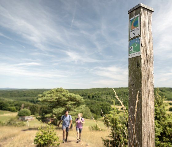 Ein Holzpfosten mit Eifelsteig-Wegmarkierung steht im Vordergrund, dahinter zwei Wanderer und eine grüne Landschaft unter blauem Himmel., © Eifel Tourismus GmbH, Dominik Ketz Ein Holzpfosten mit Eifelsteig-Wegmarkierung steht im Vordergrund, dahinter zwei Wanderer und eine grüne Landschaft unter blauem Himmel., © Eifel Tourismus GmbH, Dominik Ketz