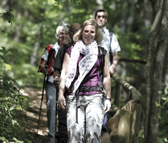 A group of hikers walk along a forest path surrounded by green trees. A woman in the foreground smiles as she walks along the path., © Wandermagazin/ N. Glatter A group of hikers walk along a forest path surrounded by green trees. A woman in the foreground smiles as she walks along the path., © Wandermagazin/ N. Glatter