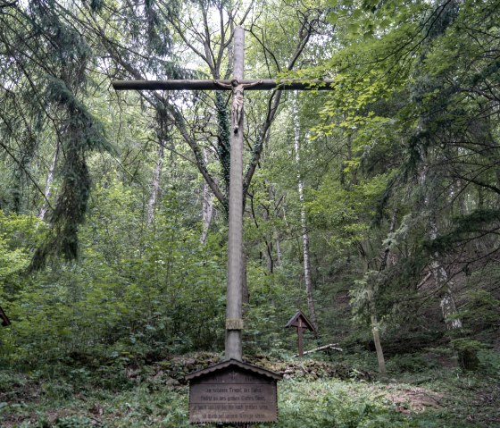 Ein großes Holzkreuz mit Jesusfigur steht in einem dichten Wald, umgeben von üppigem Grün und Bäumen., © Nordeifel Tourismus Ein großes Holzkreuz mit Jesusfigur steht in einem dichten Wald, umgeben von üppigem Grün und Bäumen., © Nordeifel Tourismus