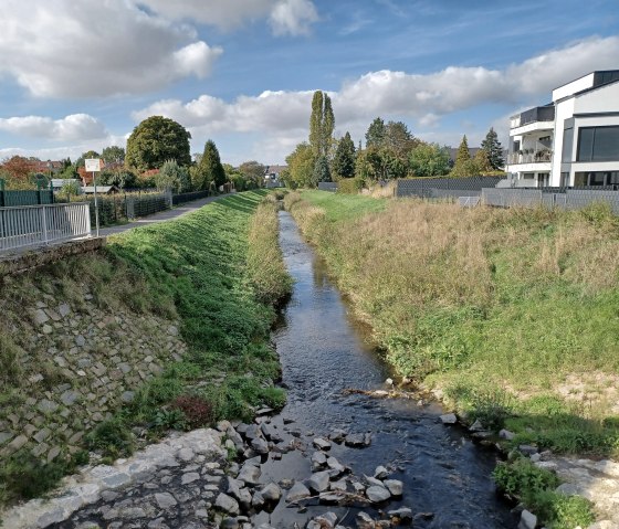 Schmaler Flussabschnitt der Swist, umgeben von grüner Vegetation, Gehweg und modernen Häusern unter blauem Himmel mit Wolken., © Sweco GmbH