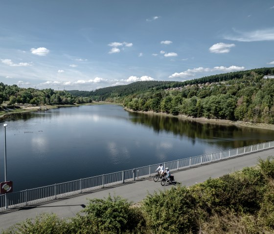 Zwei Fahrradfahrer fahren bei leicht bew&ouml;lktem Wetter auf einer Br&uuml;cke &uuml;ber den Kronenburger See. Im Hintergrund ist der Kronenburger See von einer Waldfl&auml;che umgeben, von welcher auf der rechten Seite ein Dorf herausragt., &copy; Eifel Tourismus GmbH, Dennis Stratmann