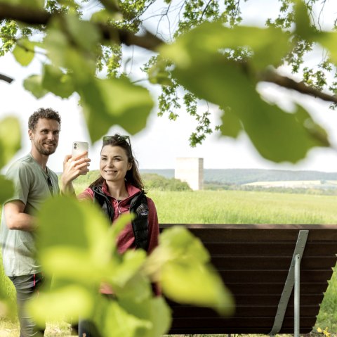 Two people take a selfie in front of the Bruder Klaus Chapel, surrounded by green leaves and a green landscape., &copy; Eifel Tourismus GmbH AR-shapefruit AG