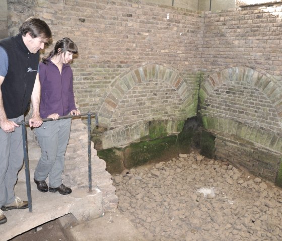 Zwei Personen stehen an einem Gel&auml;nder und betrachten eine antike r&ouml;mische Brunnenstube mit gemauerten B&ouml;gen und trockenem Boden., &copy; Wandermagazin/ N. Glatter