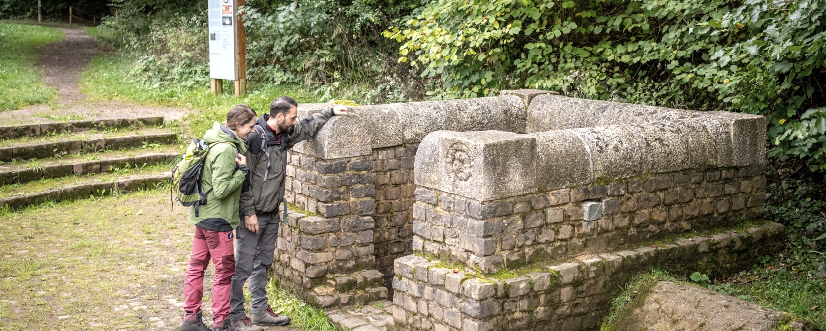 Zwei Personen betrachten einen alten Brunnen aus Stein in einem Waldgebiet. Im Hintergrund sind Bäume und ein Informationsschild zu sehen., © Eifel Tourismus GmbH, Dominik Ketz