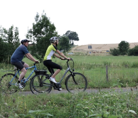 Two cyclists with helmets ride along a path through a green landscape with trees and fields in the background., © Nordeifel Tourismus GmbH