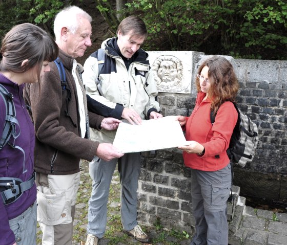 Vier Personen stehen neben einer r&ouml;mischen Quellfassung und schauen gemeinsam auf eine Karte., &copy; Wandermagazin/ N. Glatter