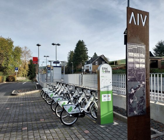 E-Bike-Verleihstation am Bahnhof Dahlem. Mehrere Fahrr&auml;der stehen bereit, daneben eine Infotafel. Im Hintergrund sind B&auml;ume und Geb&auml;ude zu sehen., &copy; Sweco GmbH