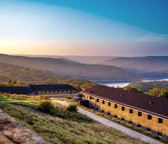 Vogelsang IP in der Eifel mit Blick auf den Urftsee bei Sonnenuntergang. Im Vordergrund historische Geb&auml;ude, im Hintergrund bewaldete H&uuml;gel und der See., &copy; Paul Meixner