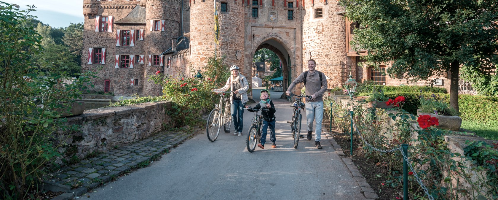 Eine Familie mit Fahrrädern steht vor der historischen Burg Satzvey. Die Burg hat markante Türme und rote Fensterläden. Es ist ein sonniger Tag., © Paul Meixner