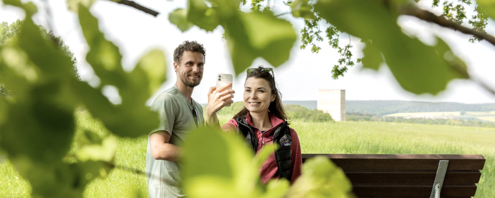 Two people take a selfie in front of the Bruder Klaus Chapel, surrounded by green leaves and a green landscape., © Eifel Tourismus GmbH AR-shapefruit AG