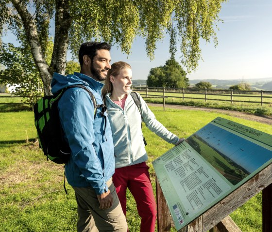 Zwei Personen stehen vor einer Informationstafel in einer gr&uuml;nen Landschaft mit B&auml;umen und einem Zaun im Hintergrund., &copy; Eifel Tourismus, Dominik Ketz