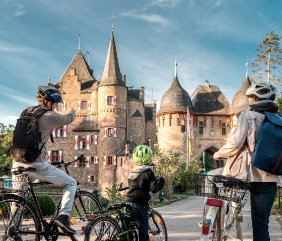 Drei Radfahrer mit Helmen betrachten die Burg Satzvey. Die mittelalterliche Burg ist von bl&uuml;henden G&auml;rten umgeben, der Himmel ist klar und blau., &copy; Paul Meixner