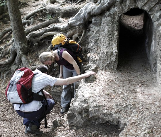 Zwei Wanderer mit Rucks&auml;cken inspizieren eine alte Wasserleitung im Wald, umgeben von Baumwurzeln., &copy; Wandermagazin/ N. Glatter