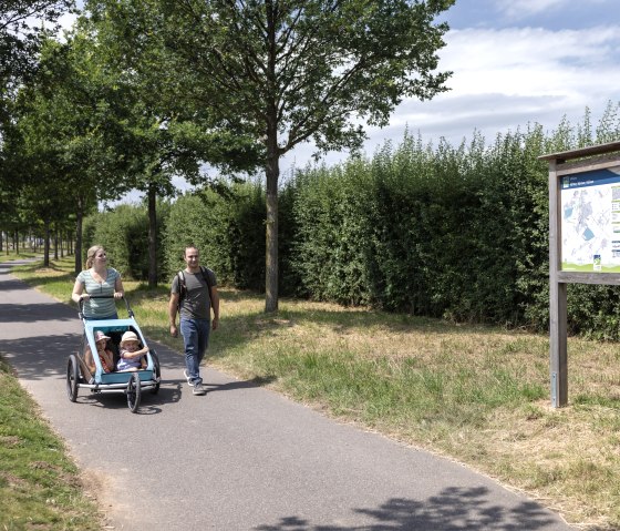 Ein Paar mit Kinderwagen spaziert auf einem asphaltierten Weg neben einer Infotafel, umgeben von Bäumen und Büschen., © Eifel Tourismus GmbH, Tobias Vollmer