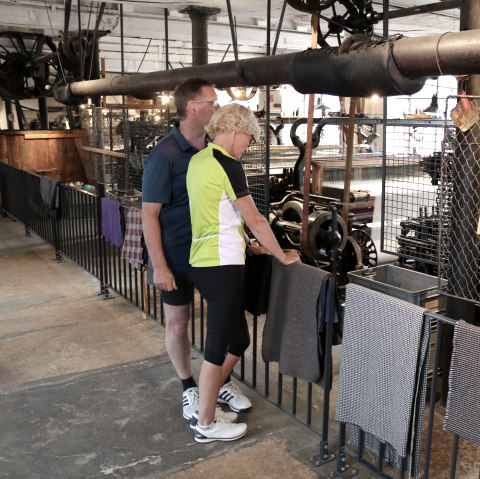 Zwei Personen stehen in einer historischen Textilfabrik und betrachten alte Maschinen. Stoffproben hängen am Geländer., © Nordeifel Tourismus GmbH