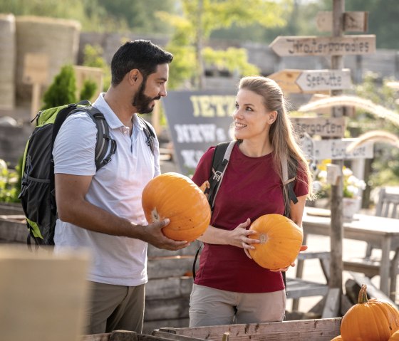Zwei Personen mit Rucksäcken halten Kürbisse auf einem Bauernhof. Im Hintergrund sind Wegweiser und Pflanzen zu sehen., © Eifel Tourismus GmbH, Dominik Ketz