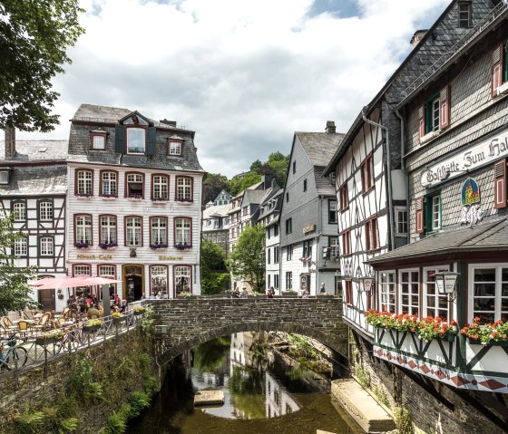 Half-timbered houses in Monschau with a stone bridge over a stream. People sit outside in a caf&eacute;. Flowers decorate the windows., &copy; Eifel-Tourismus GmbH, Dominik Ketz