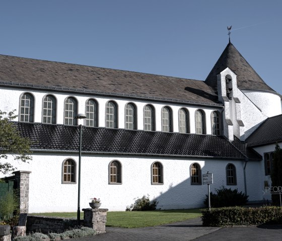 White church with round tower and pointed roof, surrounded by green spaces, under a blue sky., &copy; Nordeifel Tourismus GmbH